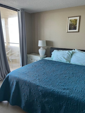 Habitación de hotel con una cama, una mesita de noche con lámpara, un cuadro en la pared y un balcón con una silla.