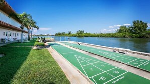 A poolside area with shuffleboard tables, a building with a white facade, and a view of a river. - The Cozy condo at Belleview Gulf (Indian Shores)