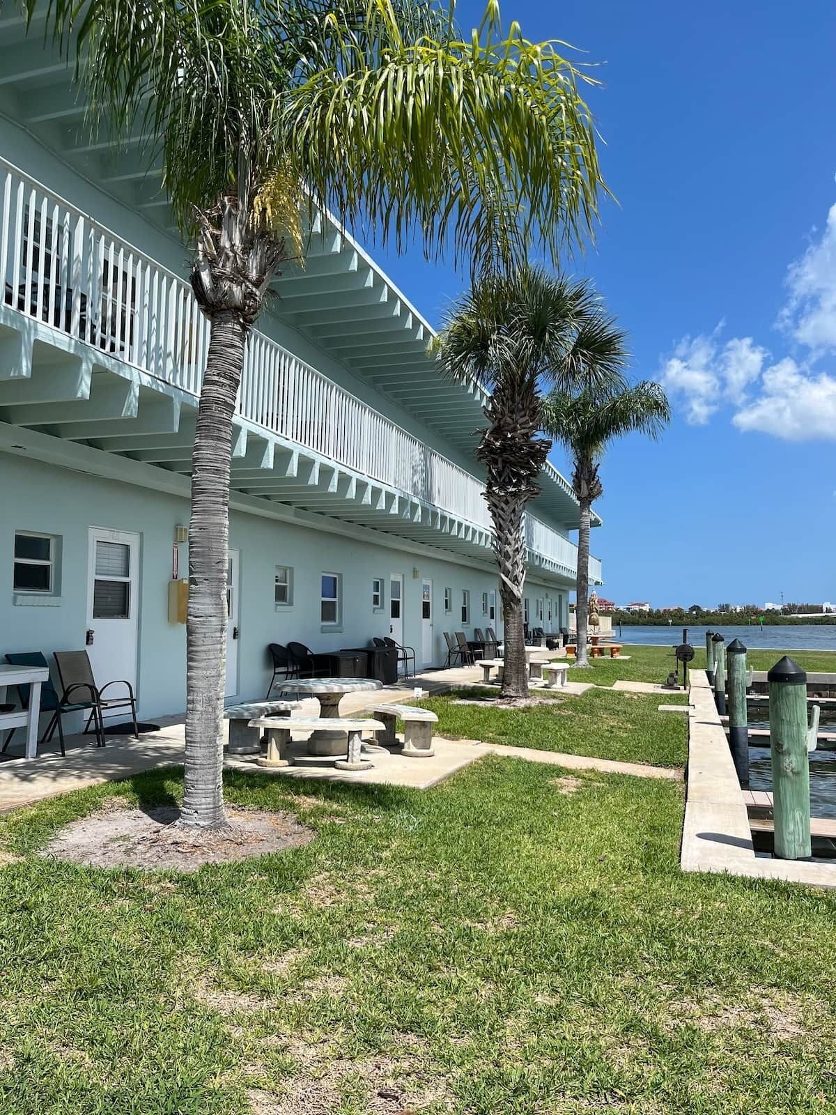 A row of white buildings with balconies, outdoor seating, and a grassy area with palm trees.