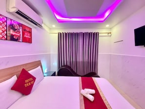 Een hotelkamer met een bed, een rood kussen met de tekst 'HOTEL TÀM MY', een televisie en een plafond met paarse verlichting.