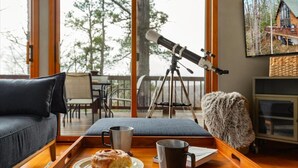 A cozy living room with a telescope on a tripod, a wooden tray with a pastry and mug, and a television displaying a cabin. - Moon View A-Frame - Wood Sauna, Mountain Views, Fire Pit, Wood Stove, Stargazing (Lost River)