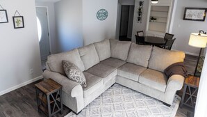 A living room with a beige sectional sofa, a small wooden side table, a patterned rug, and a dining area with a table and chairs. - The Little Blue Inn (Rochester)