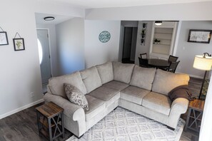 Ein Wohnzimmer mit einem beigefarbenen Ecksofa, einem kleinen Holznachttisch, einem gemusterten Teppich und einem Essbereich mit Tisch und Stühlen.