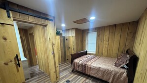 Un dormitorio con paredes y suelos de madera, una cama con ropa de cama estampada, una ventana con persianas y un ventilador de techo.
