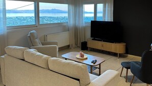 A modern living room with a sofa, a coffee table, a TV, and a view of a cityscape through large windows. - Aegean Hill Skyview (Rafina)