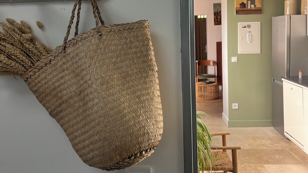 Una habitación con una cesta tejida colgada en la pared, una silla y una cocina visible a través de una puerta abierta.