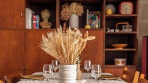 Una mesa de comedor lista con platos blancos, cubiertos y copas de vino, rodeada de sillas de madera. En el centro, un jarrón blanco con plantas secas.