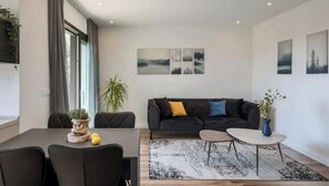 En moderne stue med en mørk sofa, et sofabord og et spiseområde med et bord og stole. Der er tre billeder på væggen og en potteplante.