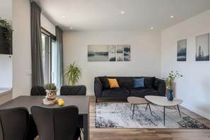Ein modernes Wohnzimmer mit einem dunklen Sofa, einem Couchtisch und einem Essbereich mit Tisch und Stühlen. Drei Bilder sind an der Wand angebracht und es steht ein Blumentopf dort.