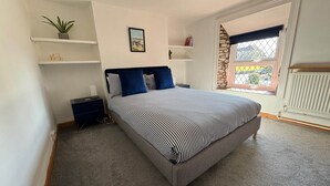 A bedroom with a bed, a nightstand, a shelf with a plant, a picture on the wall, and a window with a view. - Arcadia - 2-Bed, Dog Friendly with EV Charger (Mere)
