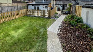 A well-maintained backyard with a wooden fence, a paved pathway, and a garden bed with mulch and plants. - Arcadia - 2-Bed, Dog Friendly with EV Charger (Mere)