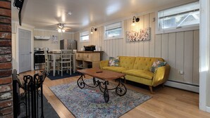 A compact living space with a yellow sofa, a wooden coffee table, a patterned rug, a dining area with a table and chairs, and a kitchen area with appliances. - Lower Unit  Killington Ski Stay w/ Fireplace & EV Charger (Killington)
