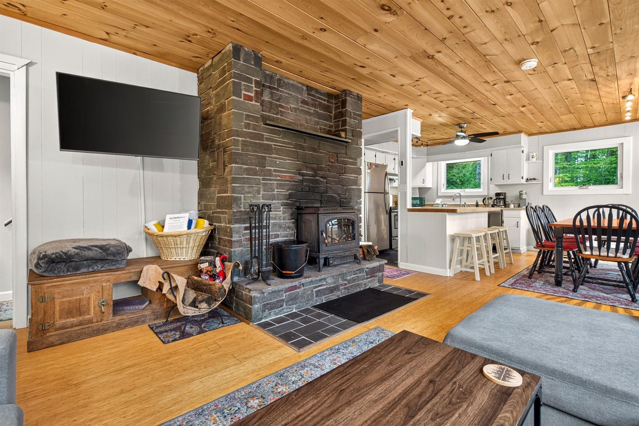 Step into your cozy open-plan retreat with warm wood ceilings, a charming wood-burning fireplace, and kitchen with modern touches. Natural light pours through windows framing lush greenery, inviting you to settle in and unwind.