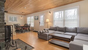 A spacious living room with a wooden ceiling, a gray sofa, a dining table with chairs, and large windows with blinds. - Vermont Mountain Escape – Spacious Duplex with Privacy & Amenities (Killington)