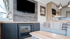 Ein modernes Wohnzimmer mit Kamin, einem flachbildfernseher, der an einer Holzwand montiert ist, einem grauen Sofa mit karierten Kissen und einer Küchenzeile mit Holzausstattung und einer Mikrowelle.