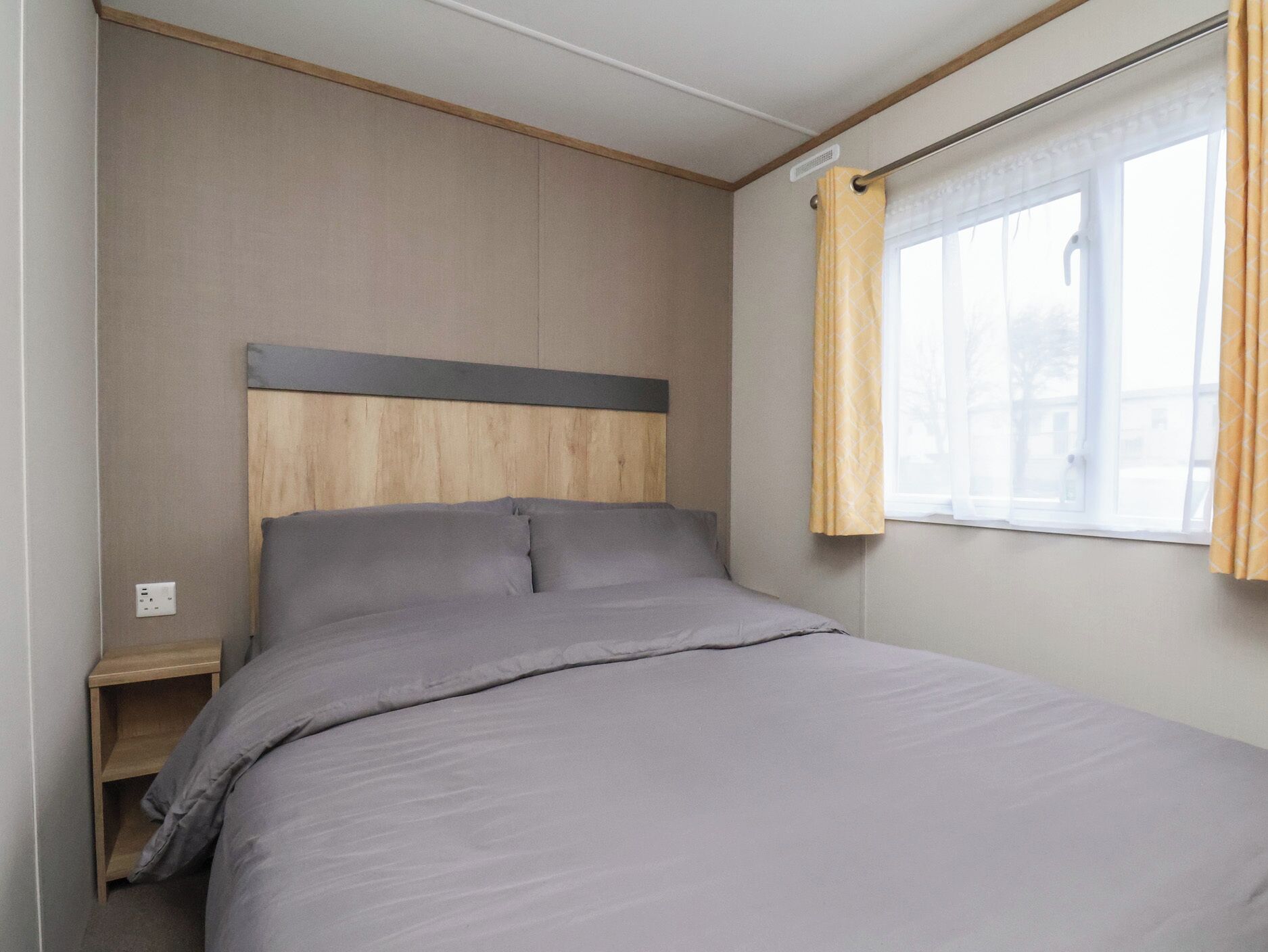A single bed with a wooden headboard in a room with a window and a power outlet.