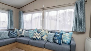 Una sala de estar acogedora con un sofá gris decorado con varios cojines estampados, ventanas amplias con cortinas translúcidas y piso de madera.