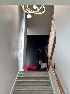 Eine Treppe mit Holzstufen und einem weißen Geländer führt in einen dunklen Raum mit einer dekorativen Leuchte und einem kleinen Tisch mit einem Schild.