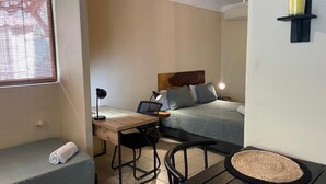 Een kleine kamer met een bed, een bureau, een stoel, een klein tafeltje en een raam met zonwering.