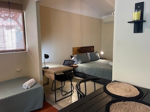 Ein kleiner Raum mit Bett, Schreibtisch, Stuhl, einem kleinen Tisch und einem Fenster mit Jalousien.