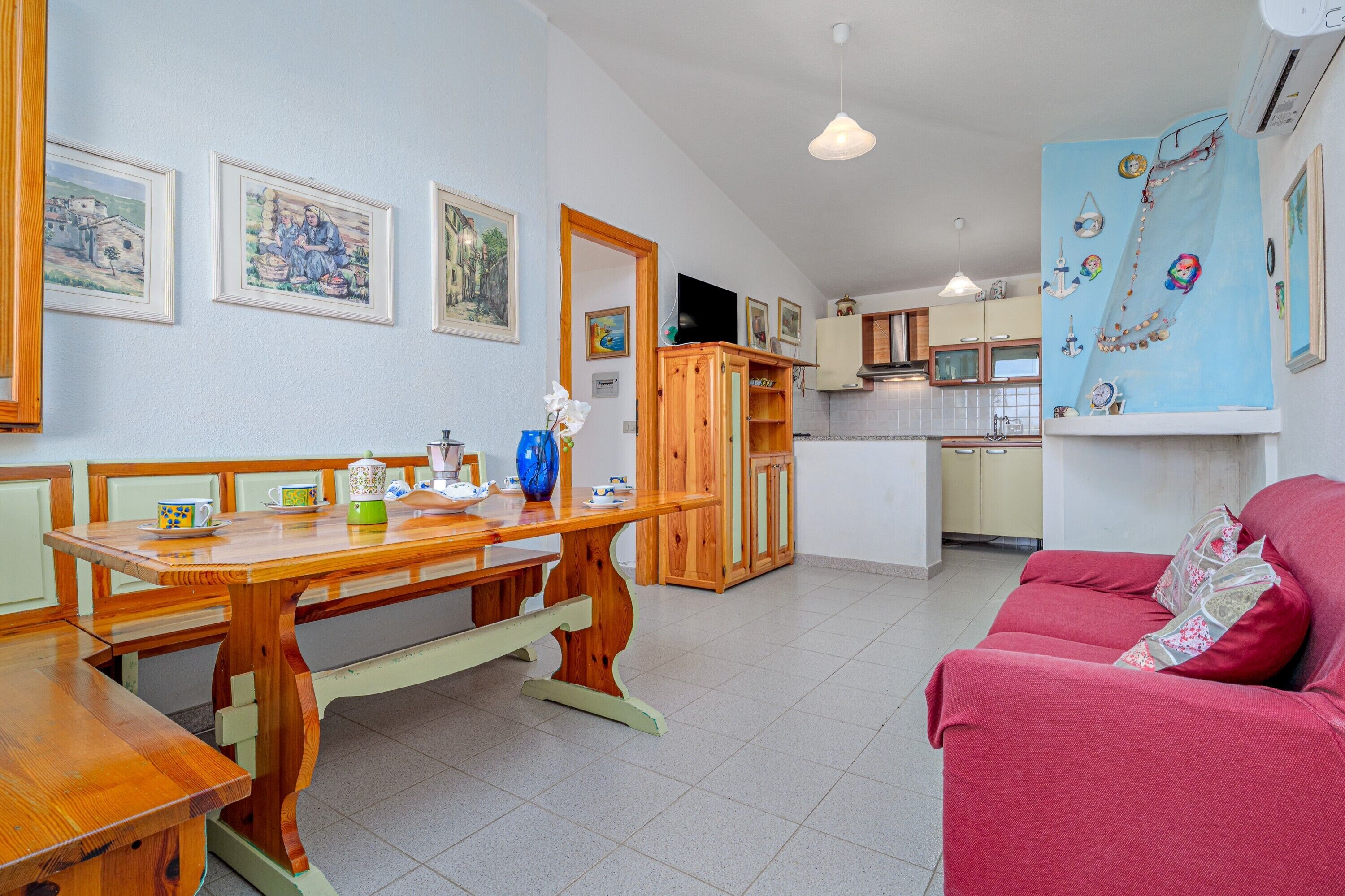A dining area with a wooden table and benches, a kitchen in the background, and a red sofa in the foreground.