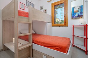 Een stapelbed met een houten ladder, een enkel bed met een oranje matras, een rode plankenwand en een raam met uitzicht op een gebouw.