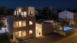 Une maison moderne à deux étages, dotée d’une piscine sur le toit, de balcons et d’un éclairage extérieur soigné pour la nuit.
