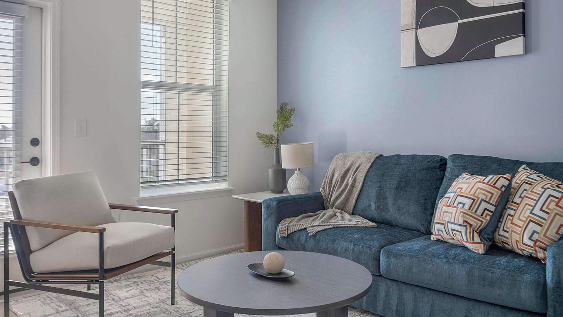 A living room with a blue sofa, a round coffee table with a decorative item, a white armchair, and a window with blinds.
