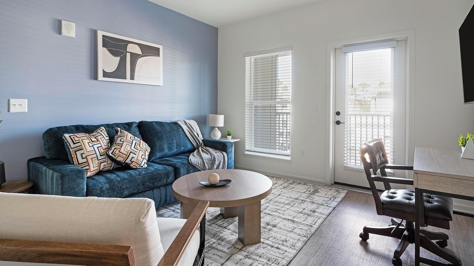 A modern living room with a blue sofa, a round coffee table, and a leather chair. There are two windows with blinds, a door, and a small table with a lamp.