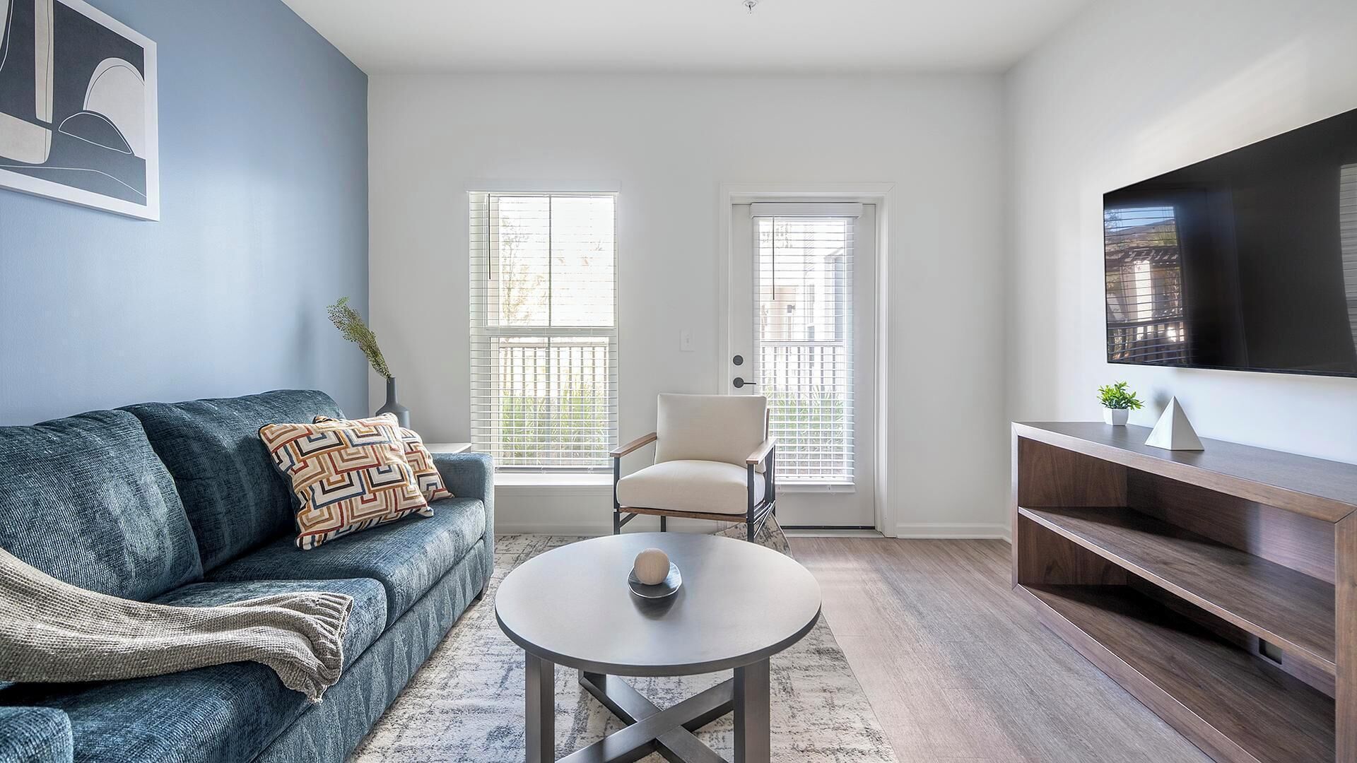 A modern living room with a blue sofa, a round coffee table, a white armchair, and a TV on a wooden stand.