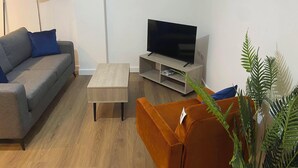 Ein Wohnzimmer mit grauem Sofa, einem kleinen Holztisch, einem Fernseher auf einem Holzschrank und einer Pflanze.