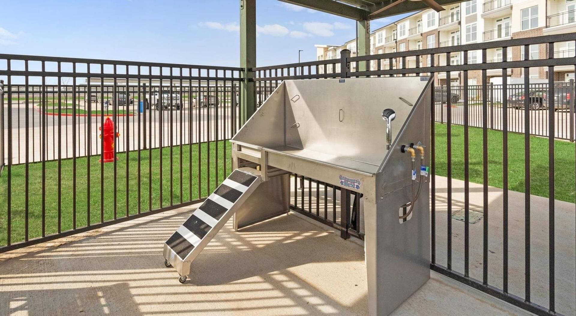 A portable water fountain with a metal ramp for wheelchair access, located in a fenced outdoor area with a parking lot and buildings in the background.