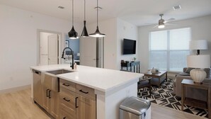 Une cuisine moderne dotée d’un îlot central, de luminaires suspendus et offrant une vue sur l’espace de vie, avec un canapé et une lampe.