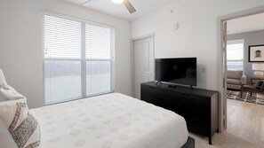 Ein Schlafzimmer mit Bett, Fernseher auf einer Kommode, einer Tür und einem Fenster mit Jalousien.