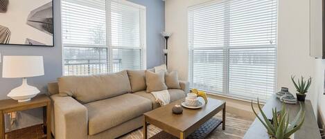 Ein Wohnzimmer mit einem beigen Sofa, einem Holztisch und einer Lampe. Große Fenster mit Jalousien, ein Teppich und ein Blumentopf sind ebenfalls vorhanden.