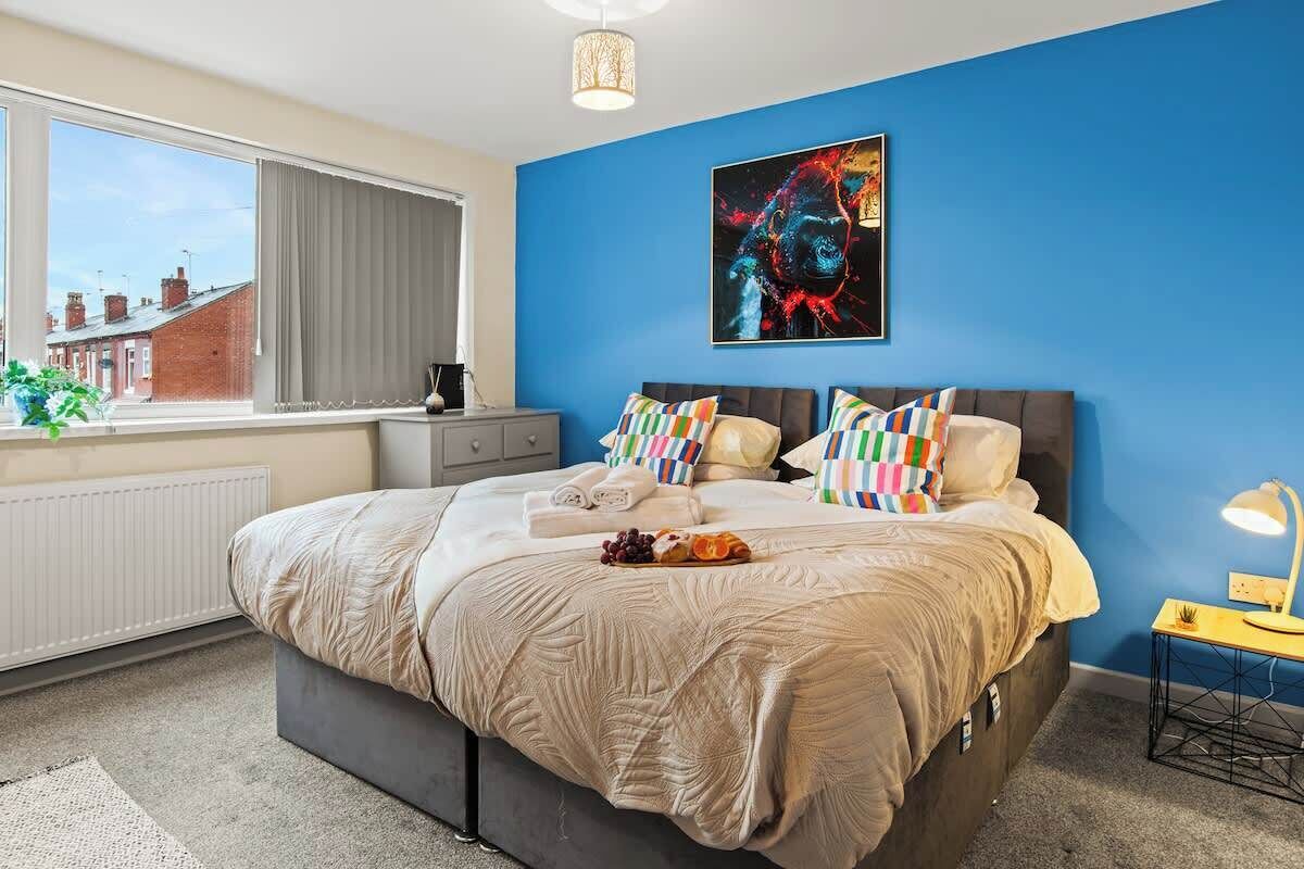 A bedroom with a large bed, a window with curtains, a radiator, a bedside table, and a wall-mounted artwork.