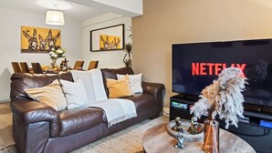 Ett vardagsrum med en brun lädersoffa, ett runt soffbord med dekorativa föremål, en platt-TV och konstverk på väggarna.