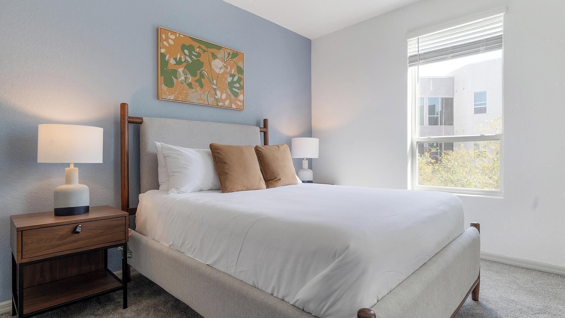 A bedroom with a bed, two bedside tables with lamps, a wall art piece, and a window with a view of a building.