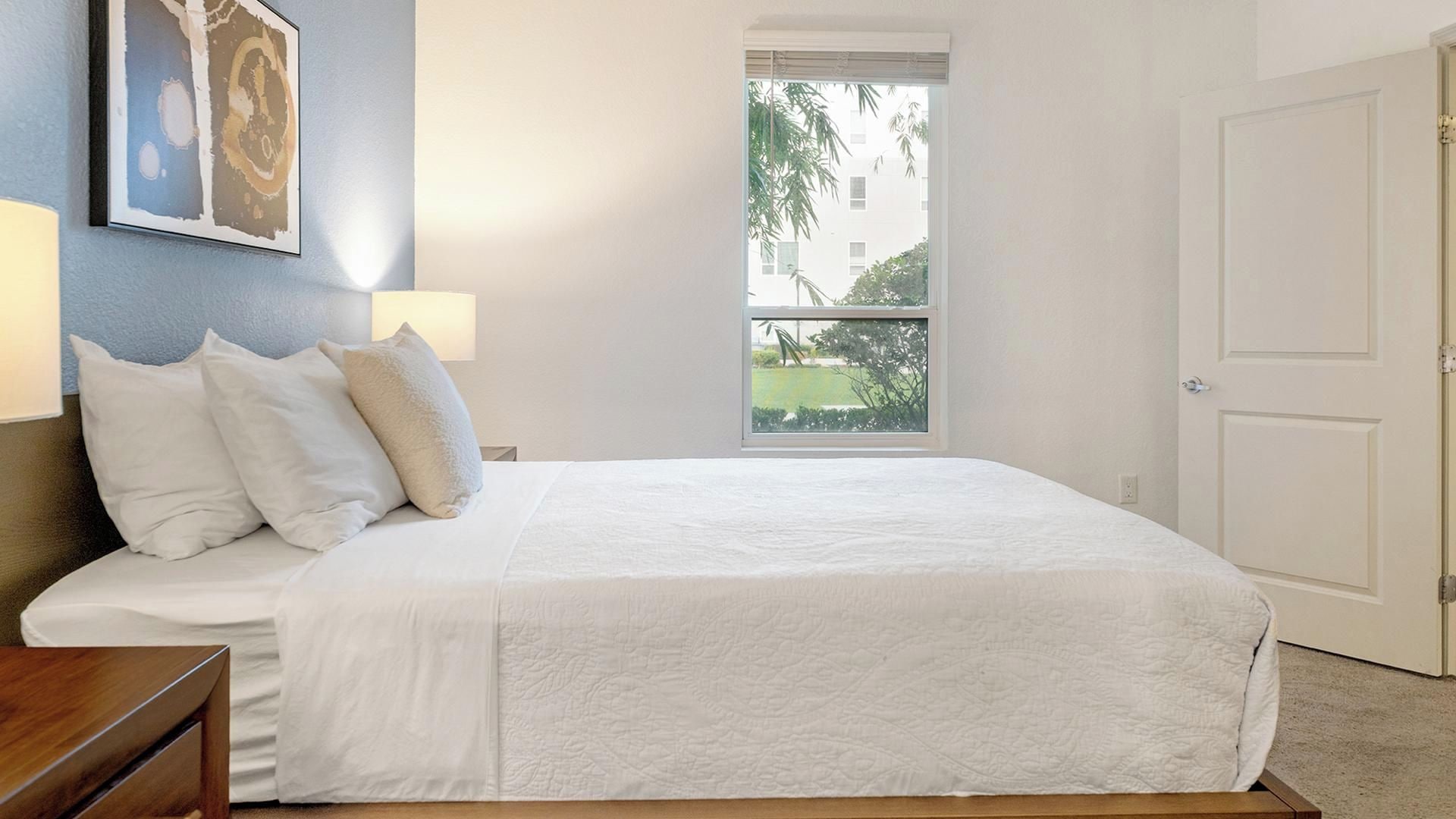 A bedroom with a bed, pillows, a nightstand, a window with a view, and a door.
