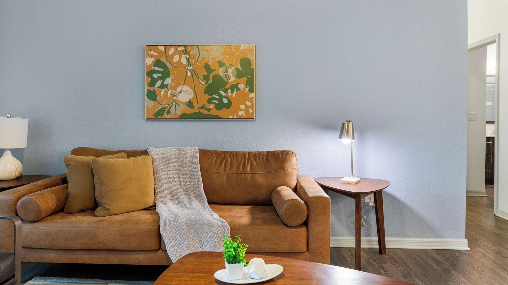 A living room with a brown leather sofa, a side table with a lamp, and a wall-mounted abstract painting.