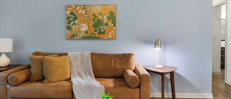 A living room with a brown leather sofa, a side table with a lamp, and a wall-mounted abstract painting.