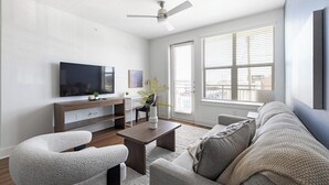Ein modernes Wohnzimmer mit einem Flachbildfernseher, Holzmöbeln, einem Deckenventilator und großen Fenstern mit Jalousien.