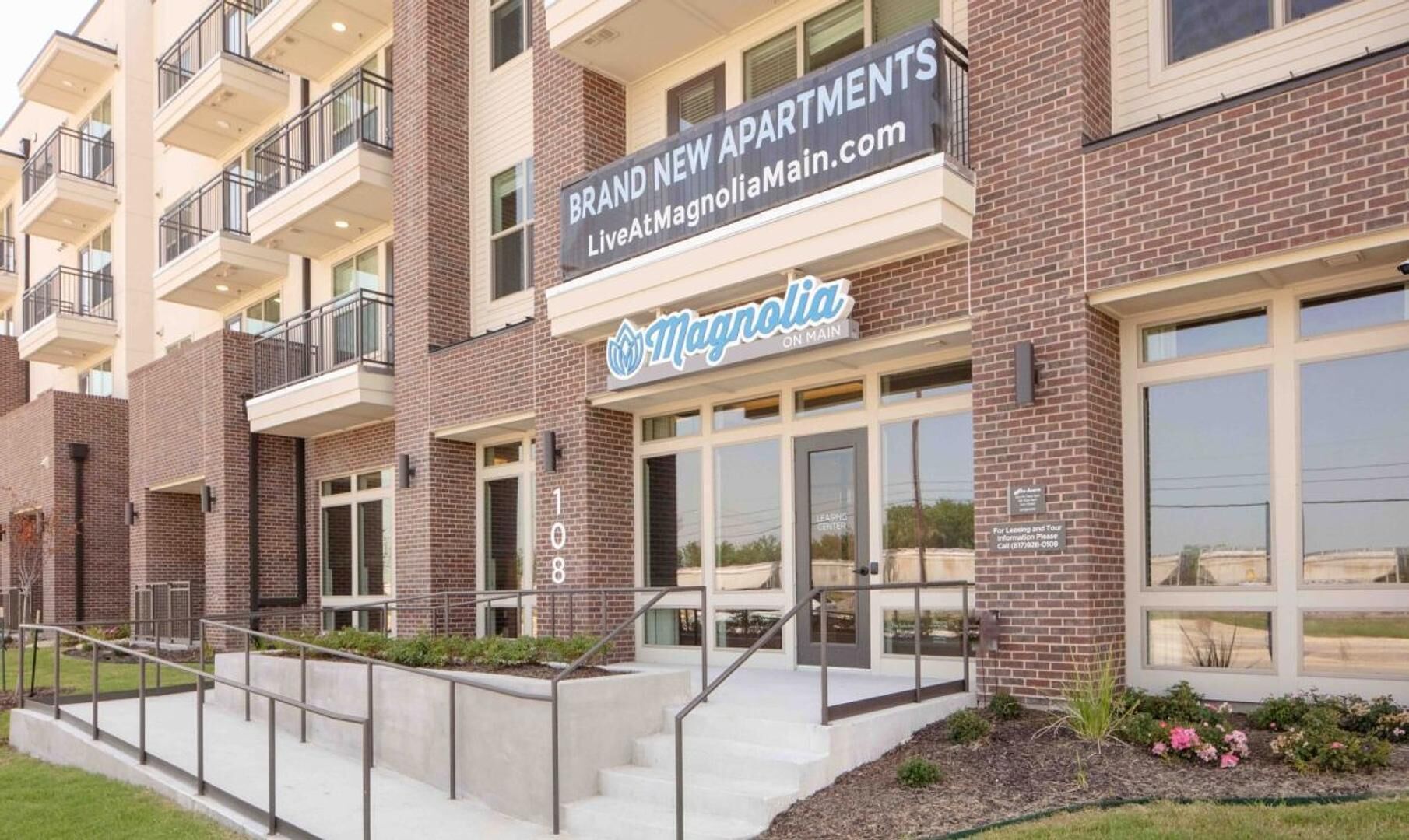 Un edificio de apartamentos de varios pisos con fachada de ladrillo, balcones y un cartel que anuncia 'Brand New Apartments' y 'Magnolia on Main'.