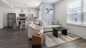 Una sala de estar moderna con un sofá gris, una mesa de centro de madera y un sillón gris. Se observa una cocina al fondo con gabinetes blancos y electrodomésticos de acero inoxidable.