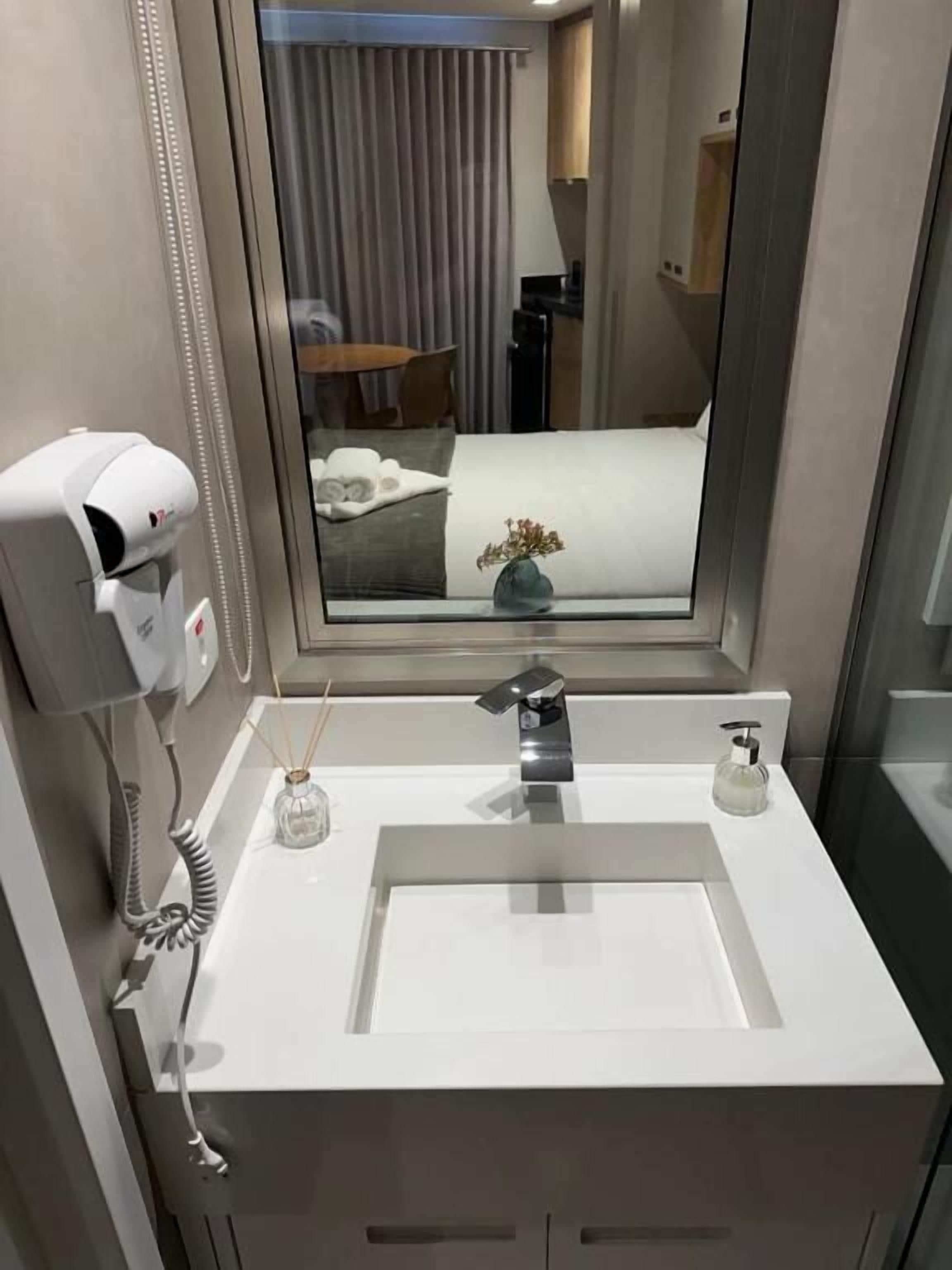 Baño de hotel con lavamanos blanco, secador de pelo, espejo y ventana con vista a una cama y una planta.