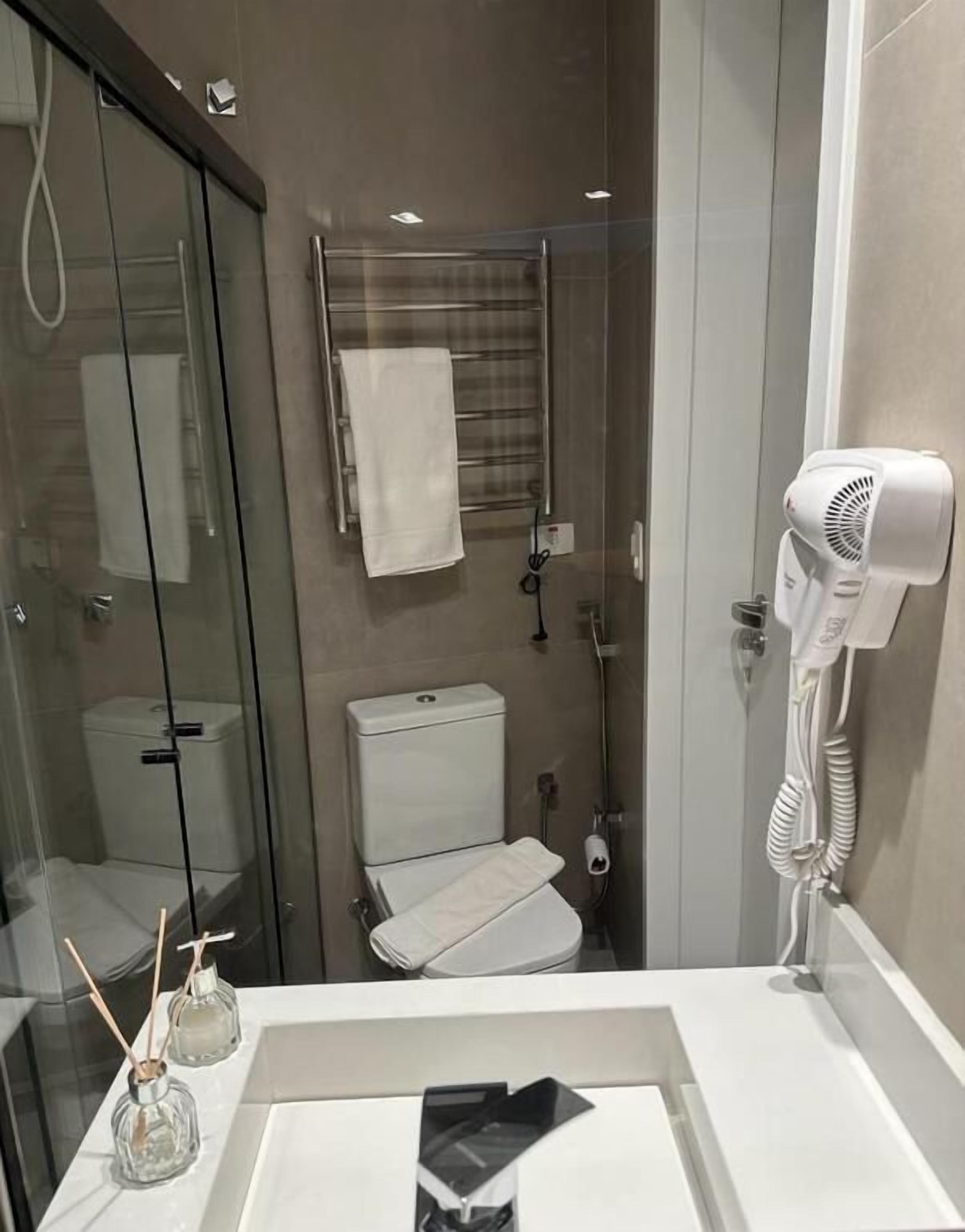 Et moderne badeværelse med brusekabine i glas, et hvidt toilet, et håndklædetørrestativ og en hårtørrer på væggen.