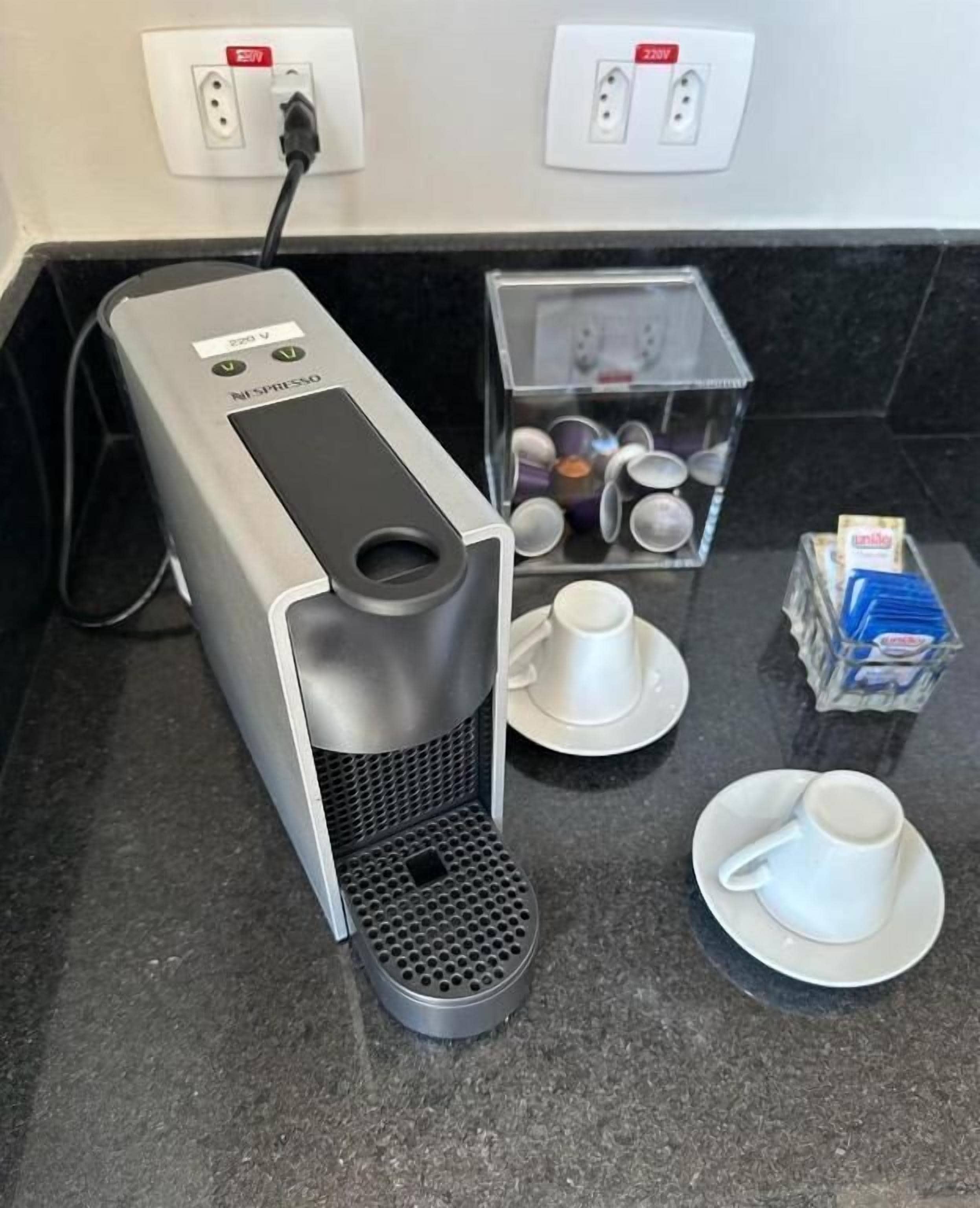 A Nespresso coffee machine with a water reservoir, a coffee pod box, and two white cups on a kitchen counter.
