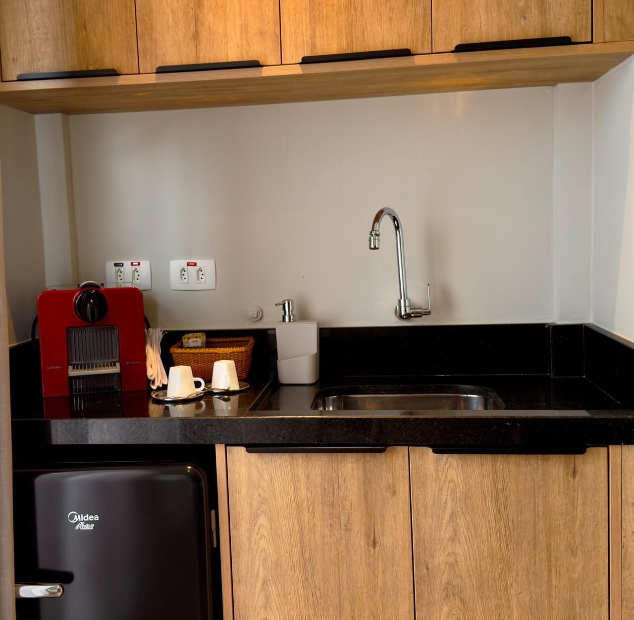 A compact kitchen with a black countertop, a stainless steel sink, a red coffee machine, and a small refrigerator.