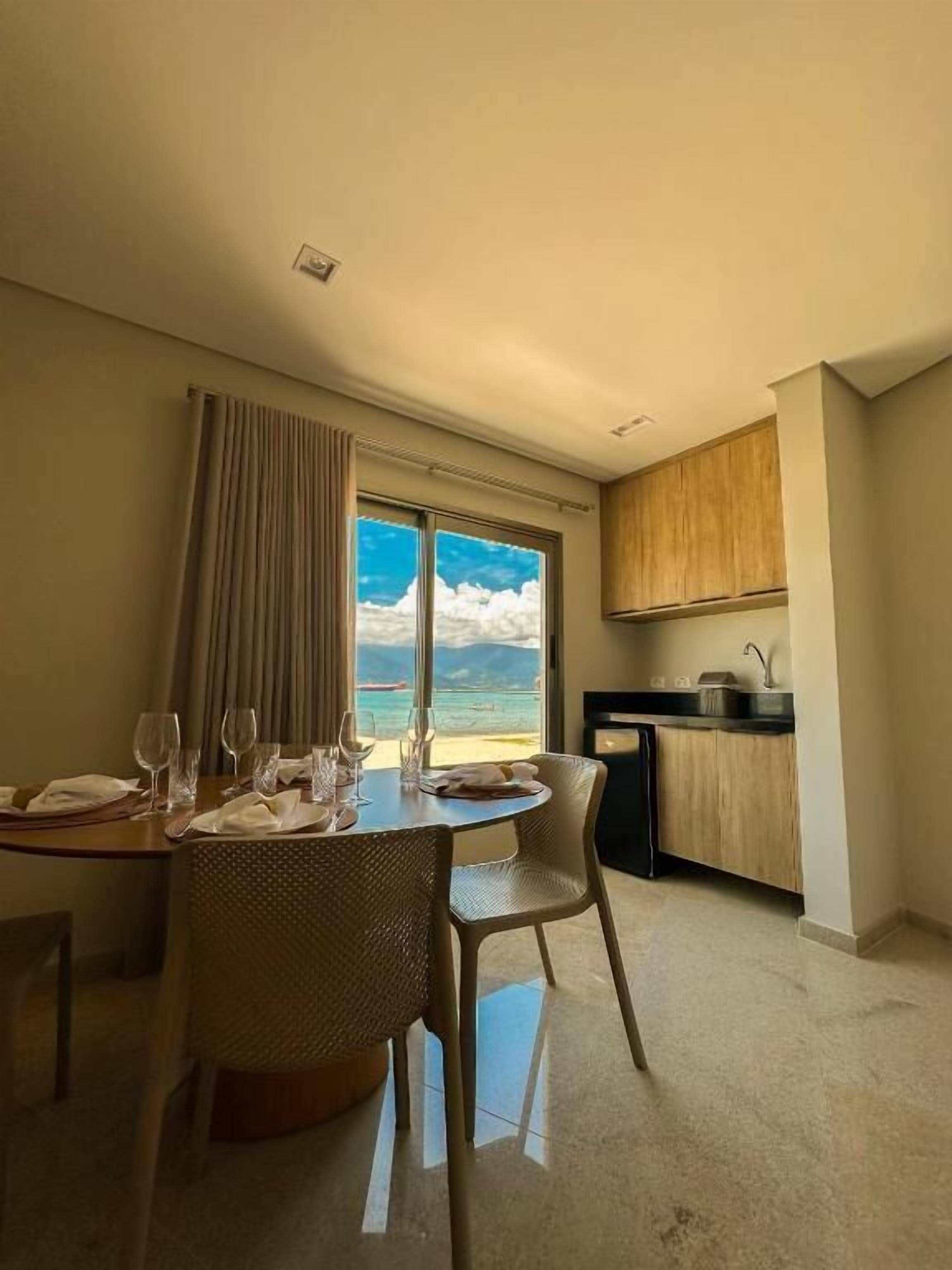 Un comedor con una mesa dispuesta para cuatro personas, vista al mar a través de amplias ventanas, y una kitchenette en la habitación contigua.