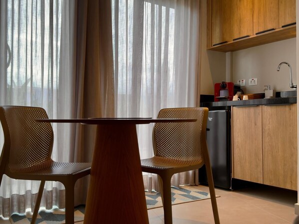 A modern kitchen with wooden cabinets, a coffee machine, and a sink. There is a dining table with two chairs in front of a window with sheer curtains. - Ágape Suítes e Apartamentos (São Sebastião)